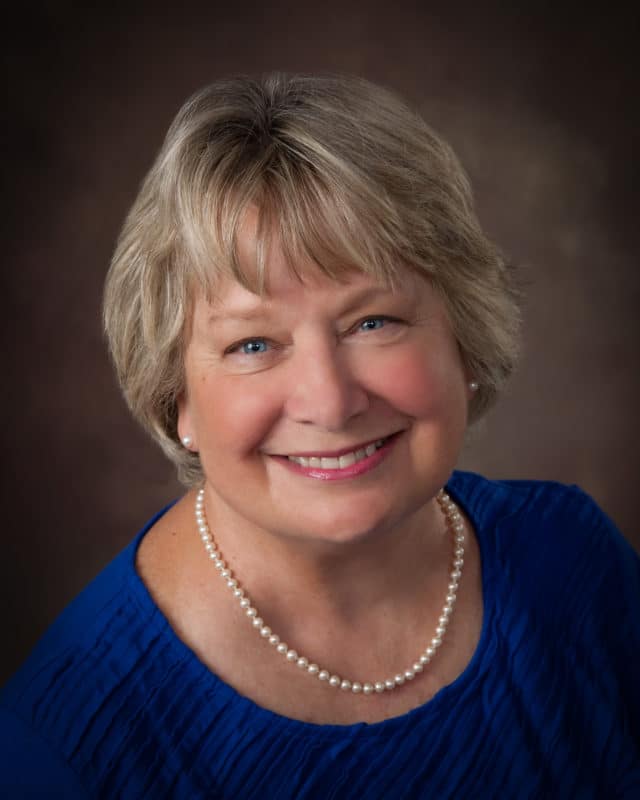 Older woman headshot wearing blue dress and pearls.