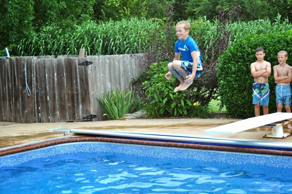 Boy in mid-air preparing to do a cannonball into a swimming pool while two friends look on.