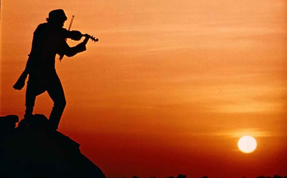 Man standing on the peak of a roof playing a fiddle, representing traditions.