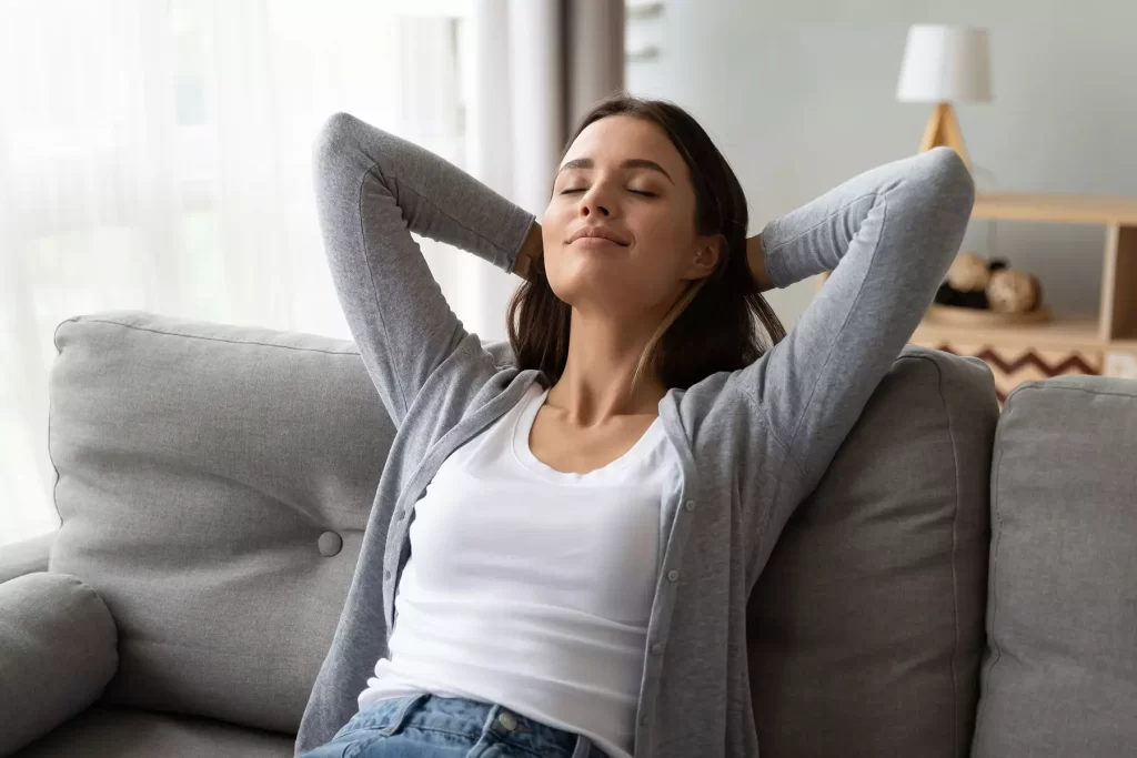 soman in grey sweater and white shirt leaning back on a sofa with her hands behind her head, relaxing.